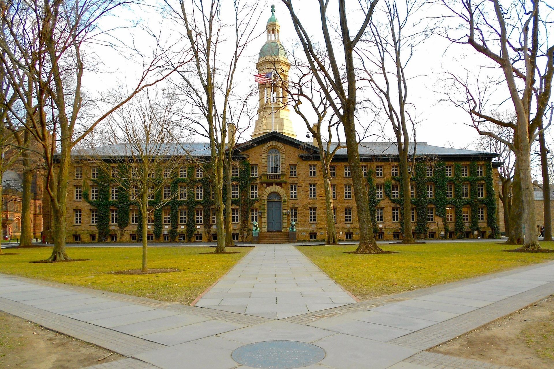 View of Nassau Hall at Princeton University, a large tan building with a gold dome and clock tower, framed by trees and walkways.