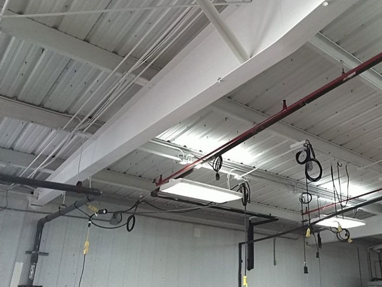 Interior view of a warehouse ceiling with metal beams, pipes, wiring, and fluorescent lights. The space is mostly white and gray.