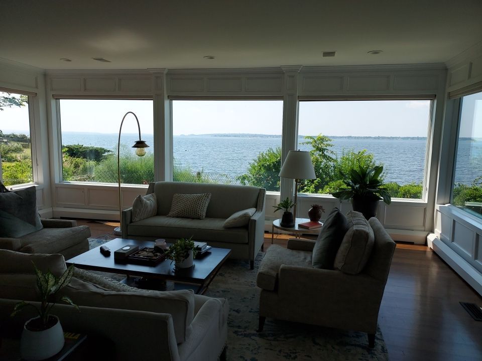 A living room with large windows offering a scenic view of a body of water. Beige furniture sits on a wooden floor, with a coffee table in the center.