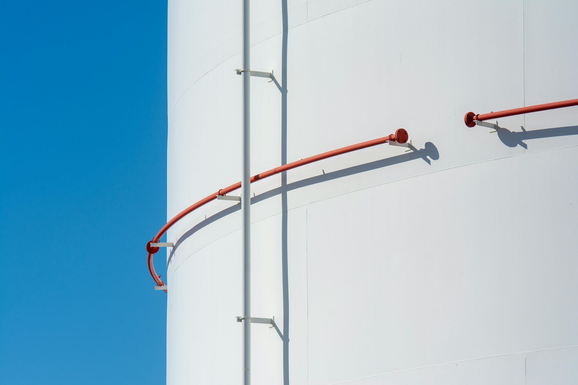 White industrial tank with a red pipe and ladder against a clear blue sky.