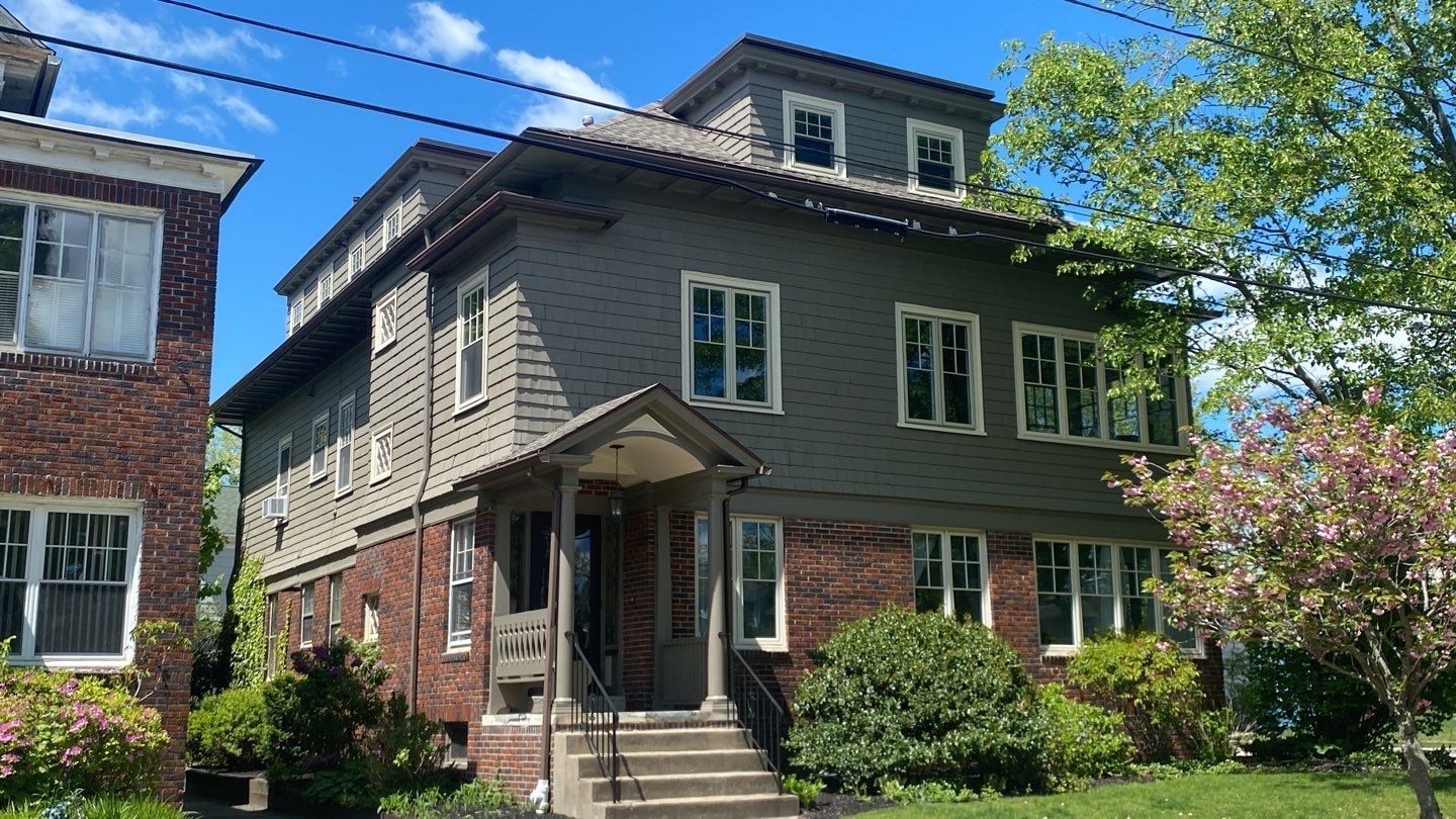 Three-story apartment building with a brick base and gray siding. A porch has stairs leading up to the entrance.