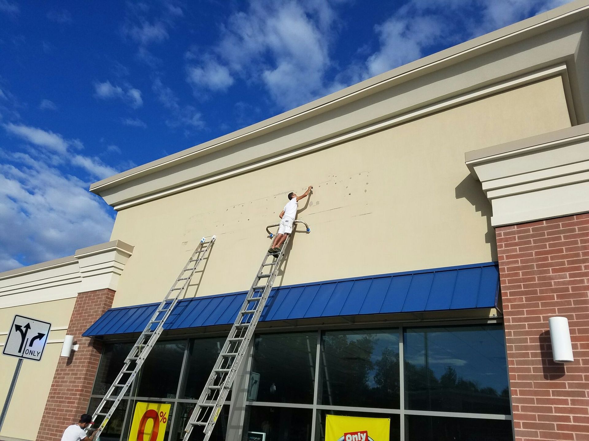 Two people painting the exterior of a commercial building. One person on a ladder is painting the tan wall, under a blue awning and blue sky.