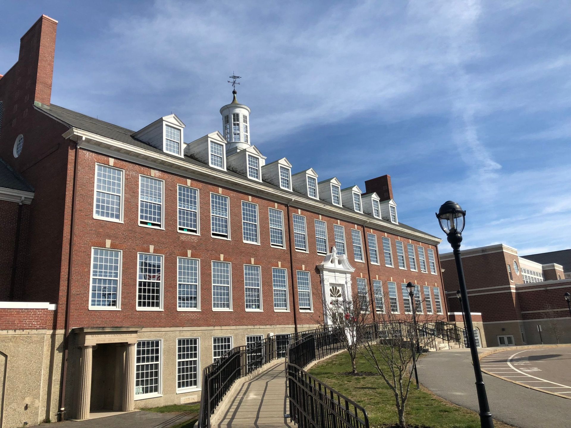 Brick building with many windows, a small cupola, and a walkway in front, under a blue sky.