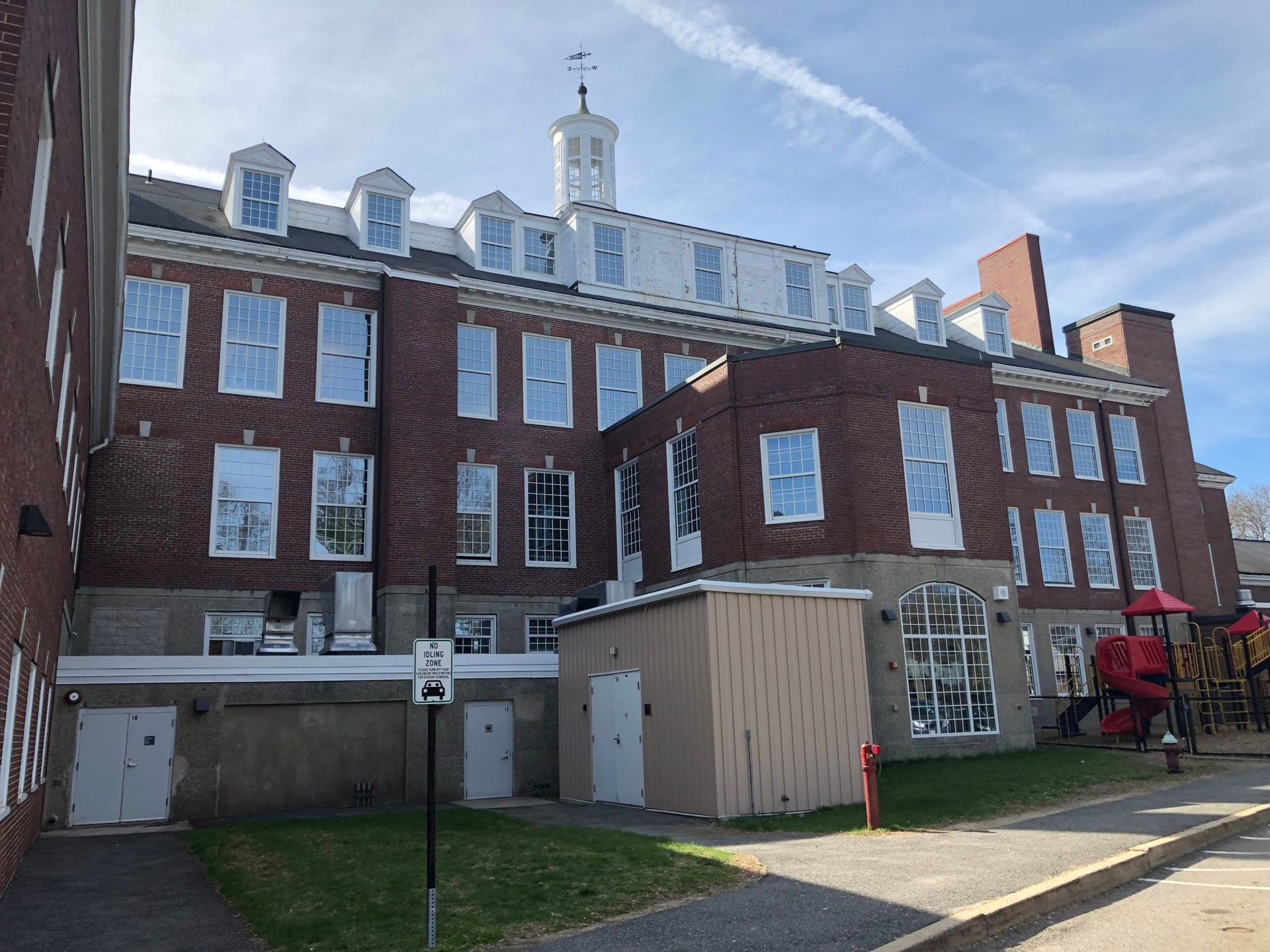 Brick building with many windows, a small tower, and a playground to the right. Sunny day.
