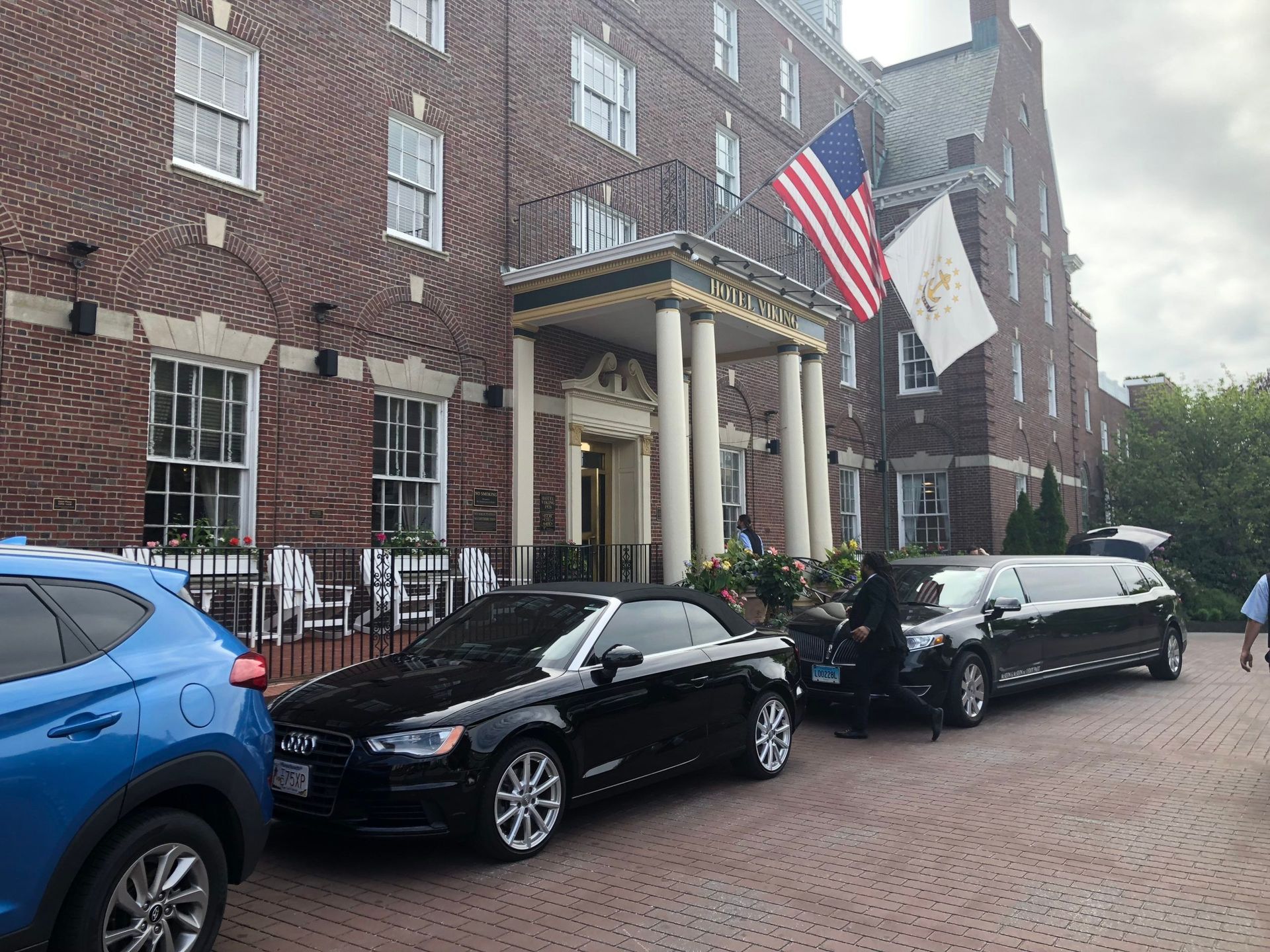 A brick building with a decorated entrance. A blue SUV, black convertible, and black limousine are parked outside. American and state flags fly above.