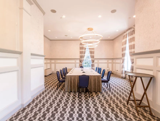 A conference room with a long table, blue chairs, patterned floor, and a decorative chandelier. There is a high-top table to the side.