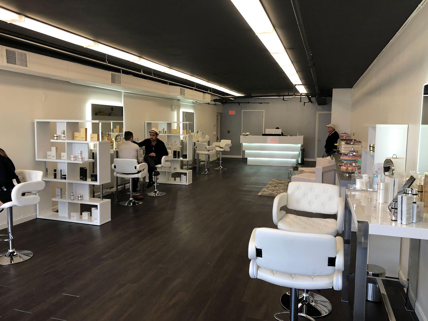 Interior view of a modern salon with white styling stations, dark wood floors, and clients receiving services. Brightly lit with white and gray decor.