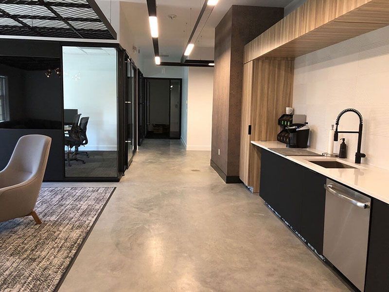 Modern office space with a kitchenette and private glass offices along a hallway. Concrete floor, black cabinets, and warm wood accents.