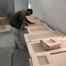 Man wearing green sweater and paint-splattered pants working on wooden cabinet doors laid out on a table in a workshop.