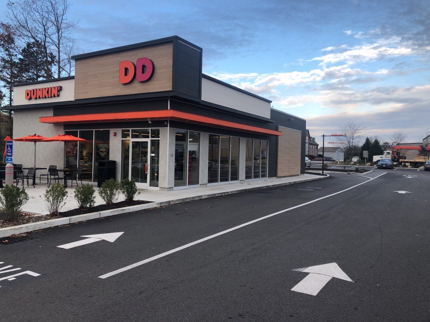 Exterior view of a Dunkin' Donuts shop with a drive-through lane marked with arrows. The building has orange and black accents and outdoor seating.