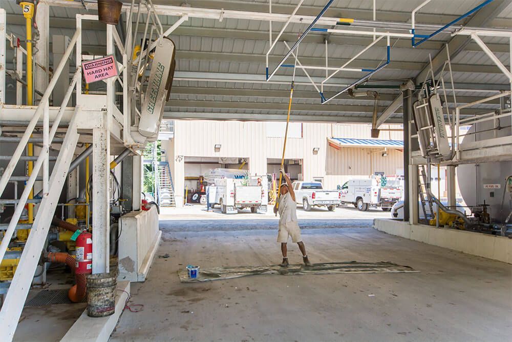 Person using a long-handled tool to clean a concrete floor in an industrial setting with vehicles parked in the background.