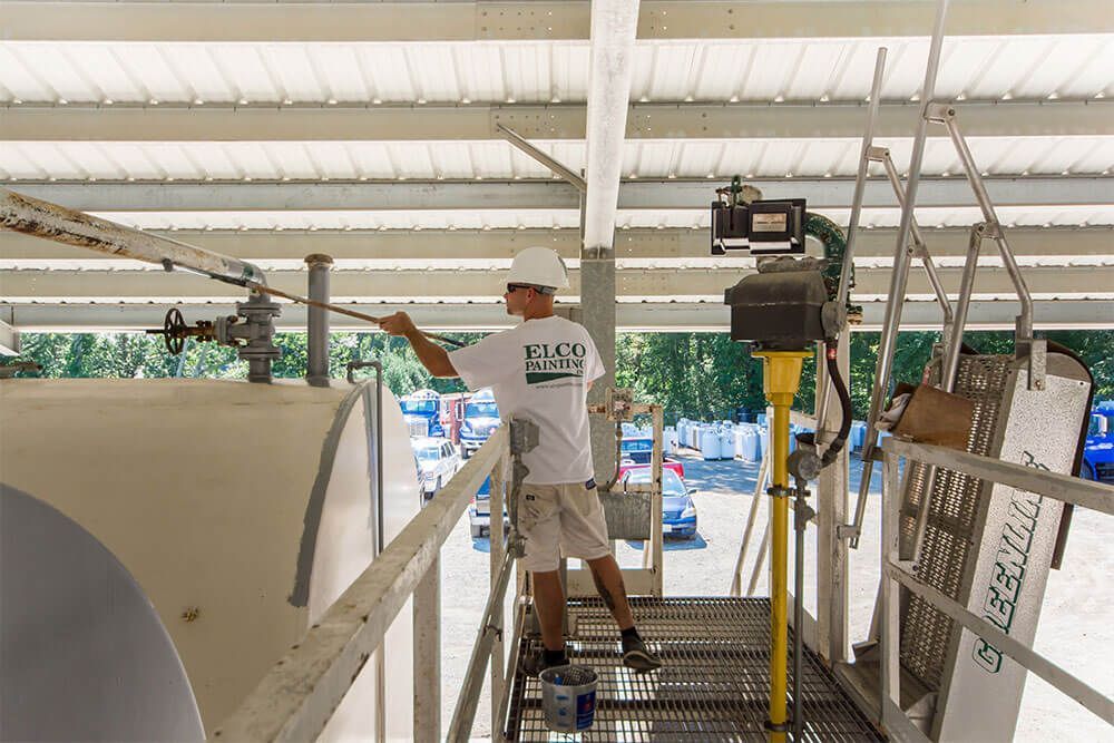 A worker in a white hard hat paints a large tank on a metal walkway. The worker is wearing a white shirt that says 