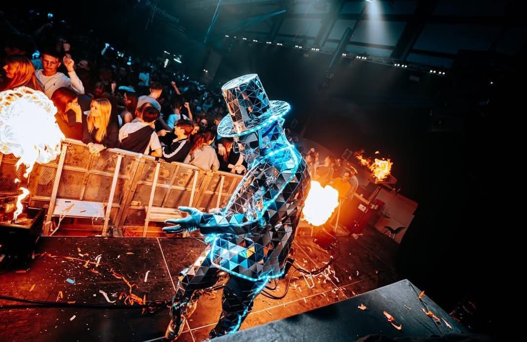 Artiest in een glanzend pak en hoge hoed met blauwe lichten, op het podium met vuurwerk. Publiek op de achtergrond.