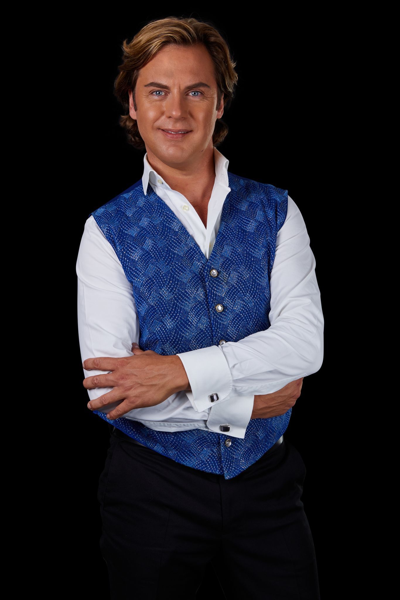 Een man in een blauw vest met patroon, een wit overhemd en een zwarte broek, met de armen over elkaar, lachend tegen een zwarte achtergrond.