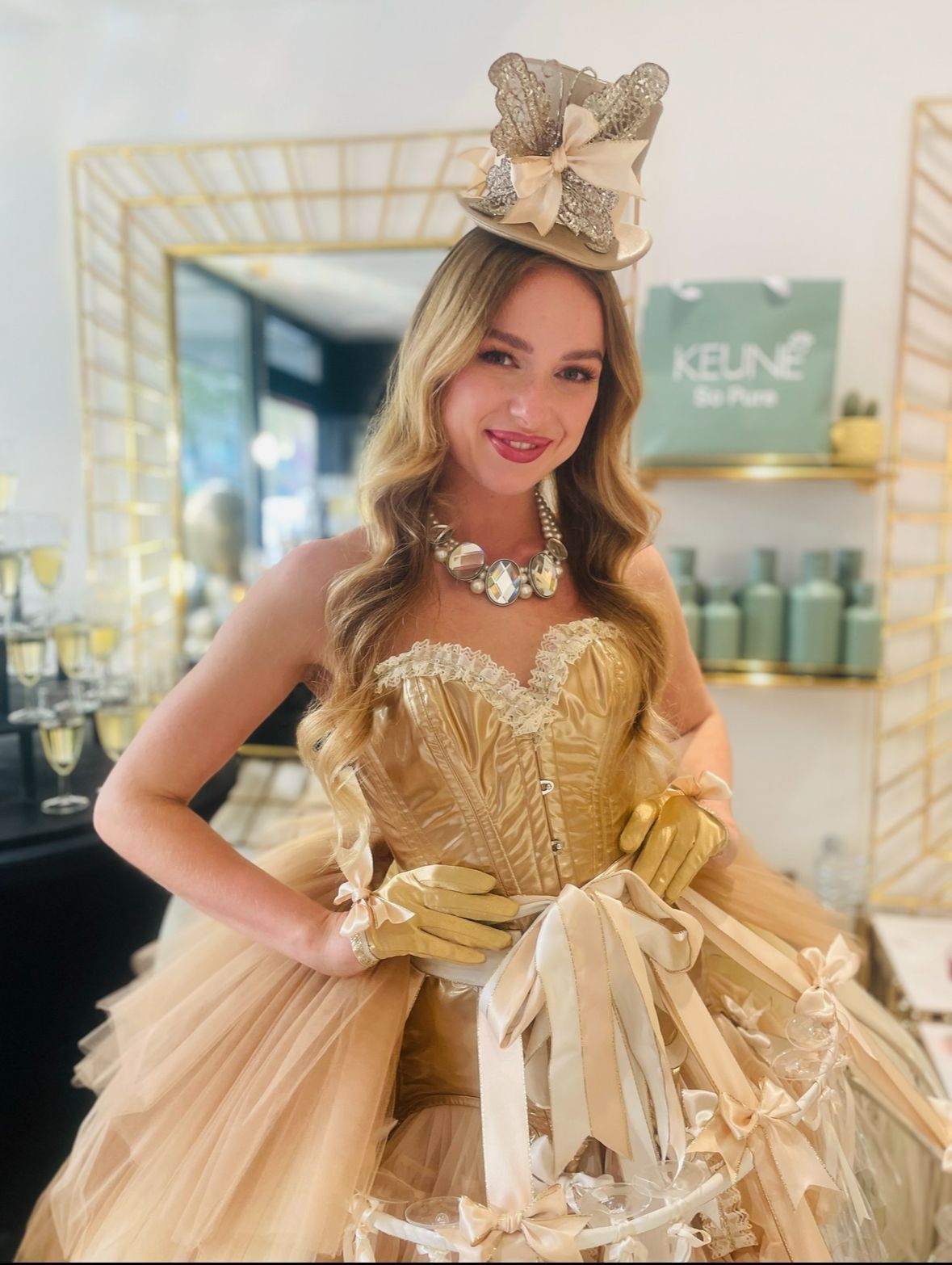 Een vrouw in een gouden jurk en hoge hoed glimlacht, staand in een salon met champagne en verzorgingsproducten op de achtergrond.