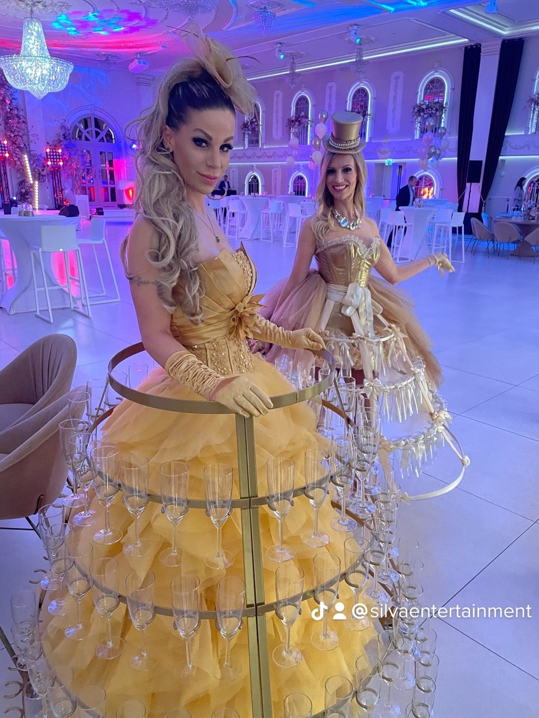 Twee vrouwen in champagnekleurige jurken, de ene geel, de andere goud, glimlachen op een formeel evenement.