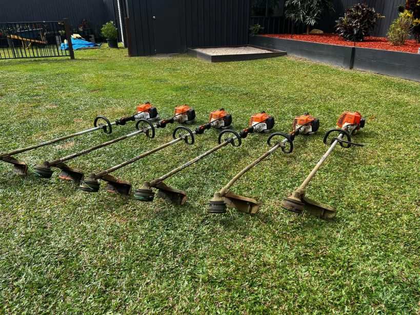 Seven String Trimmers on a Grassy Lawn, Lined Up in Front of a Building — Mow & Mulch Lawn & Garden Maintenance In White Rock, QLD