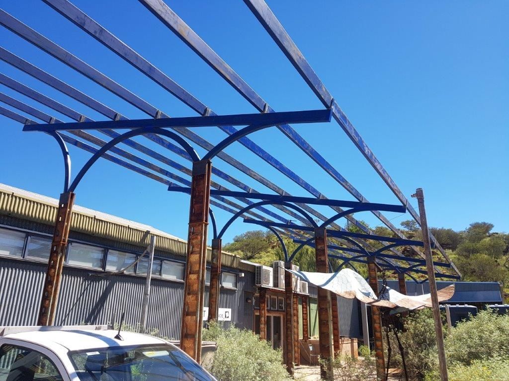 A car is parked under a metal structure in front of a building.