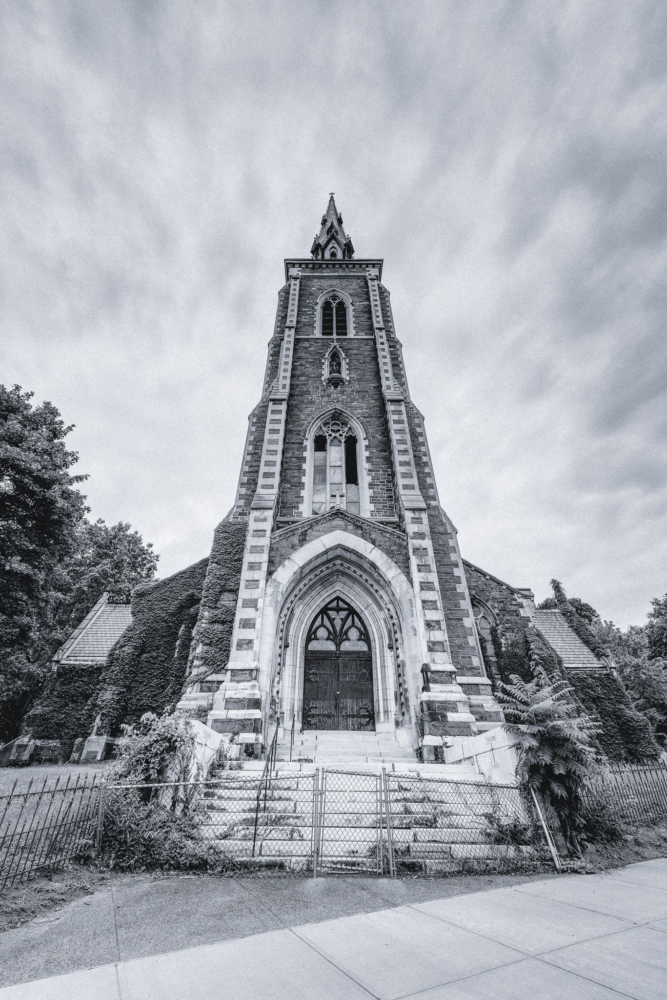 Stone tower with Gothic arch entrance, steps, and spire, under cloudy sky.