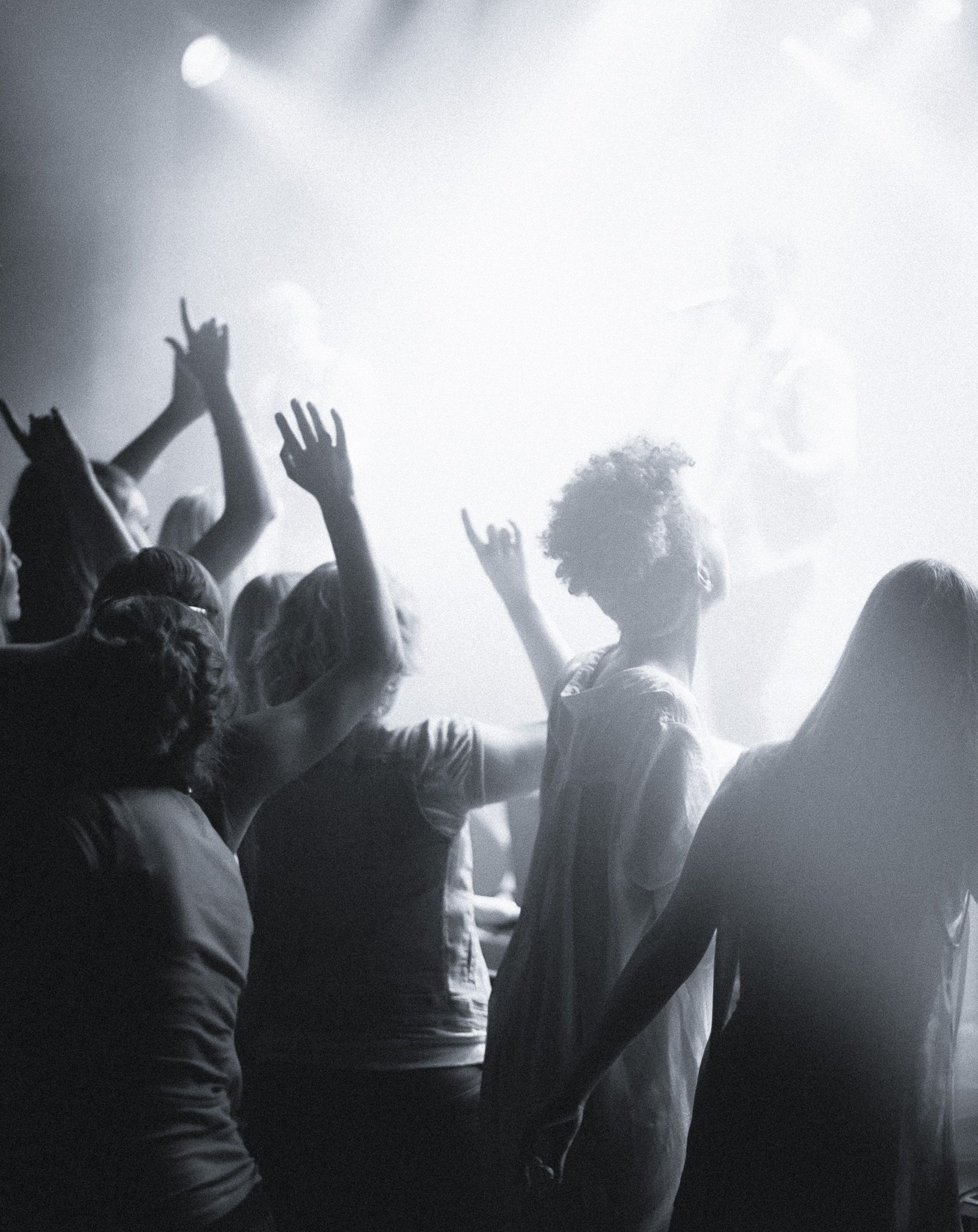 Worship concert crowd with arms raised in the air, bathed in bright stage lights.