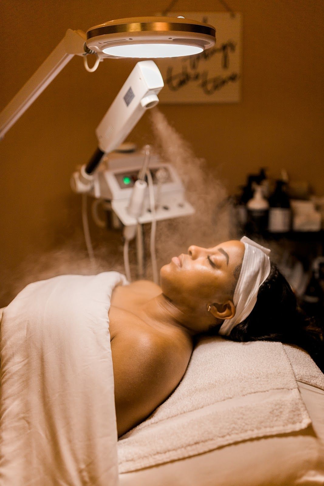 A person lies on a spa bed receiving a facial treatment with a warm steam vaporizer and professional lighting.