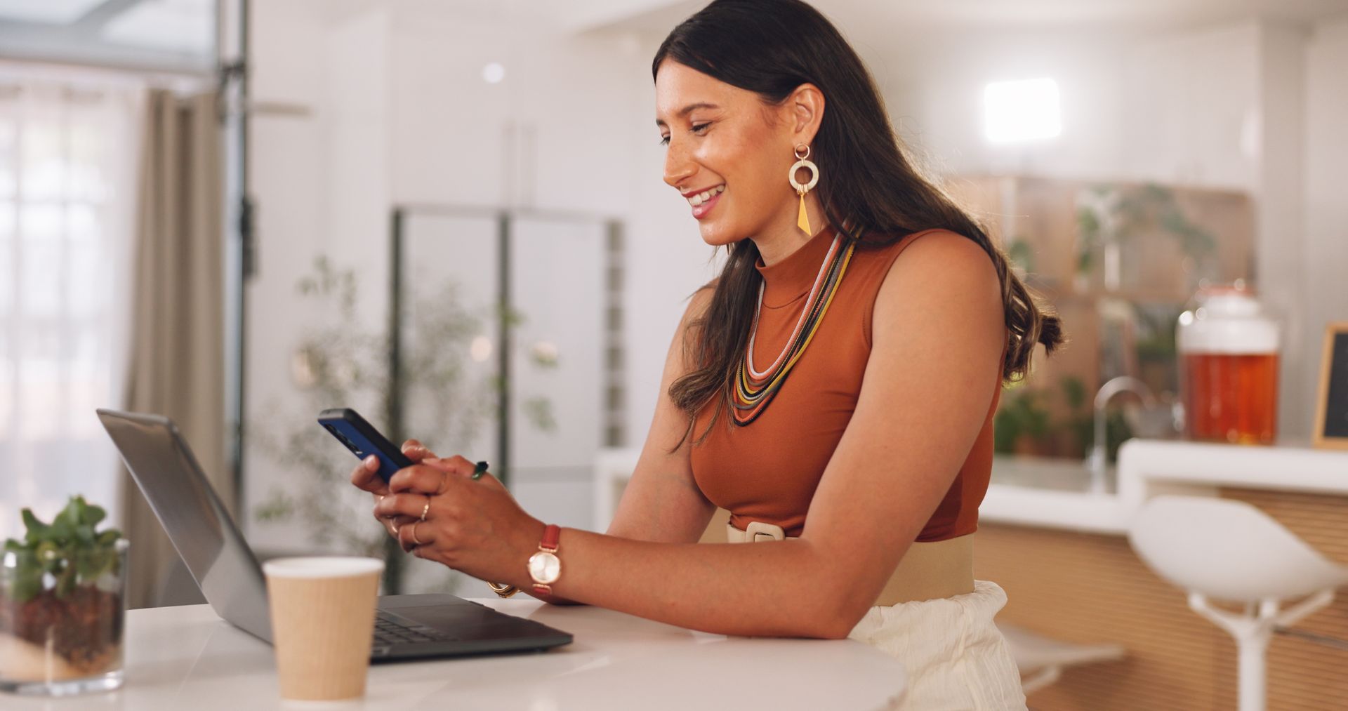 A woman is sitting at a table using a laptop and a cell phone.