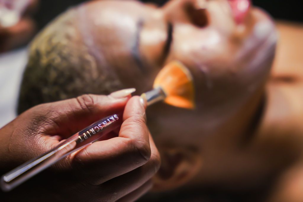 Person receiving a facial treatment; a hand holding a brush applies product to the face.