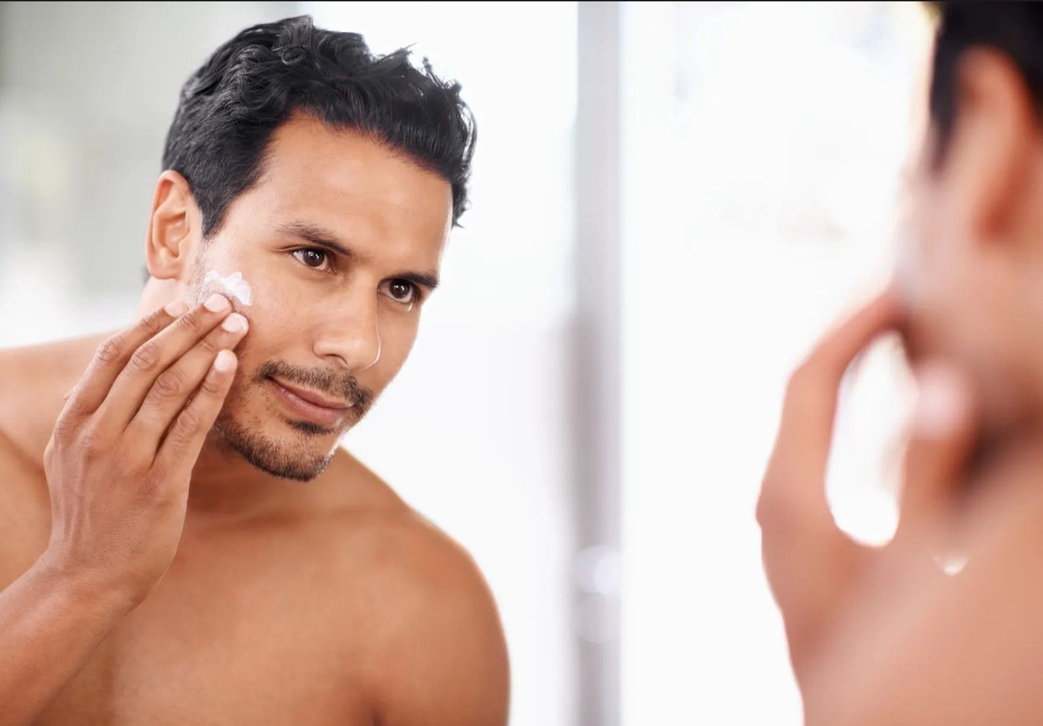 A shirtless man is applying lotion to his face in front of a mirror.