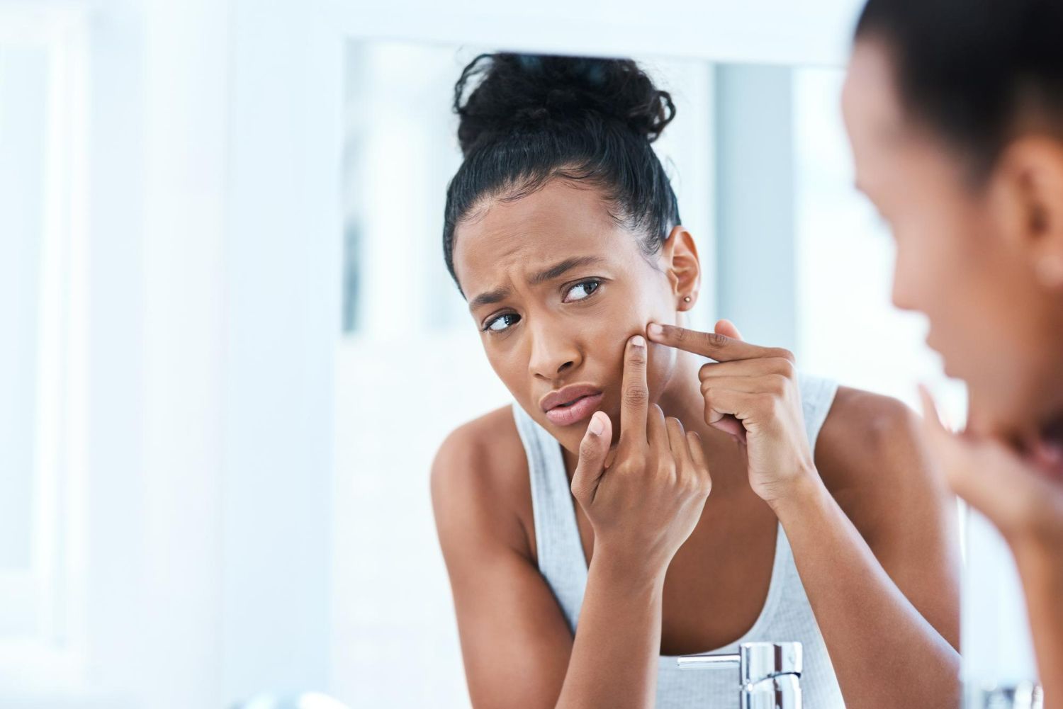 Woman with dark skin looking in mirror, inspecting a pimple on her cheek.