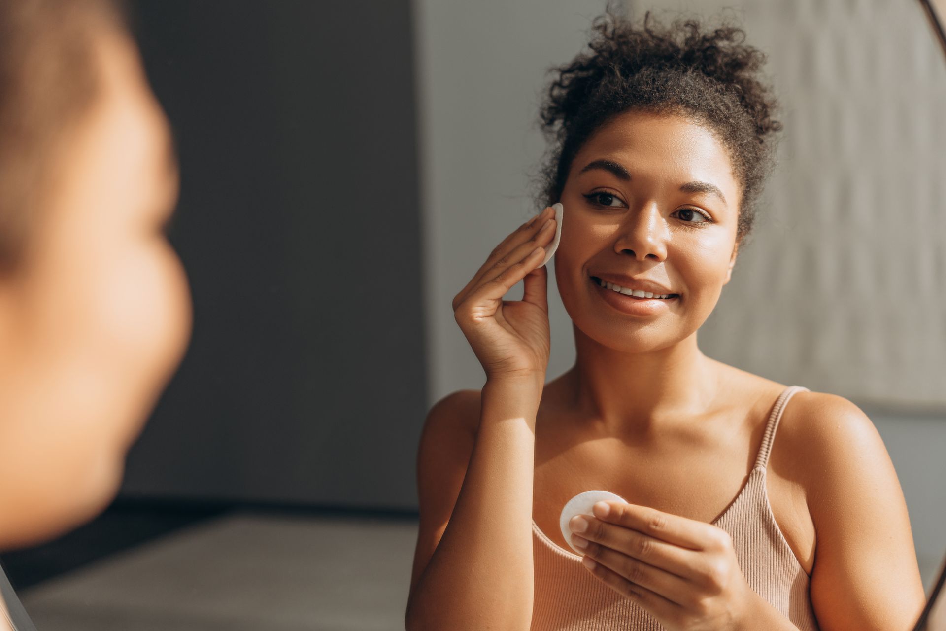 Woman uses a cotton pad on her face in a mirror, smiling, wearing a light pink top, indoors.