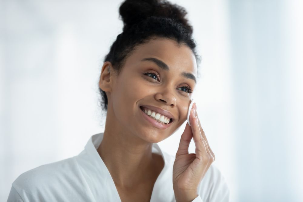 A person in a white robe smiling while applying skincare to their cheek with a cotton pad.