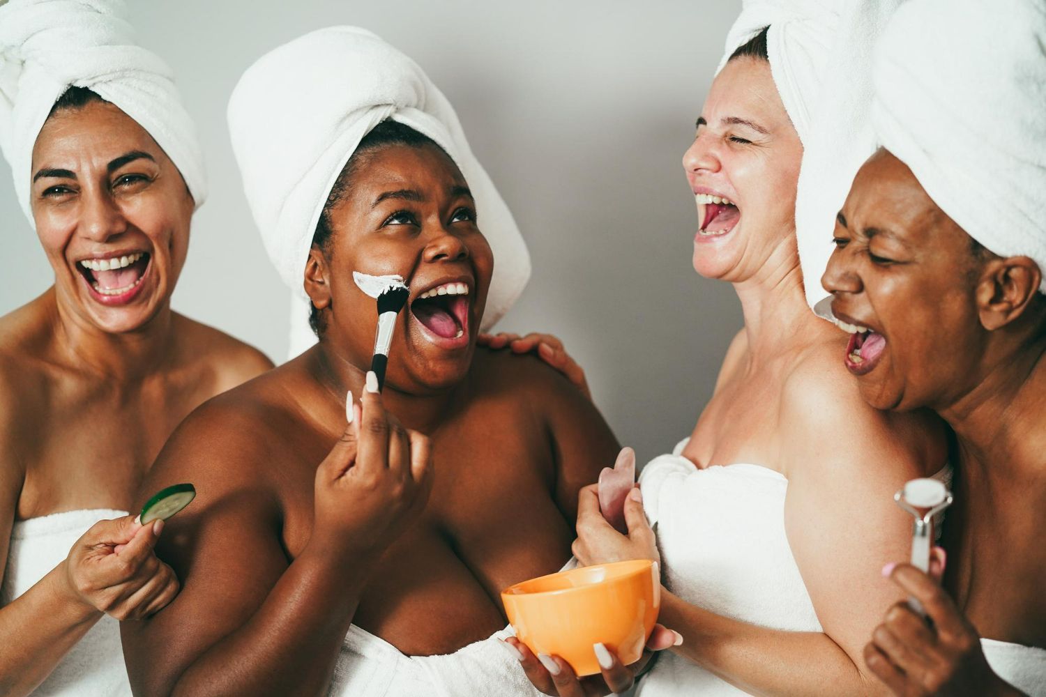 Four people wearing white head towels and towels wrapped around them laugh together while using skincare tools and masks.