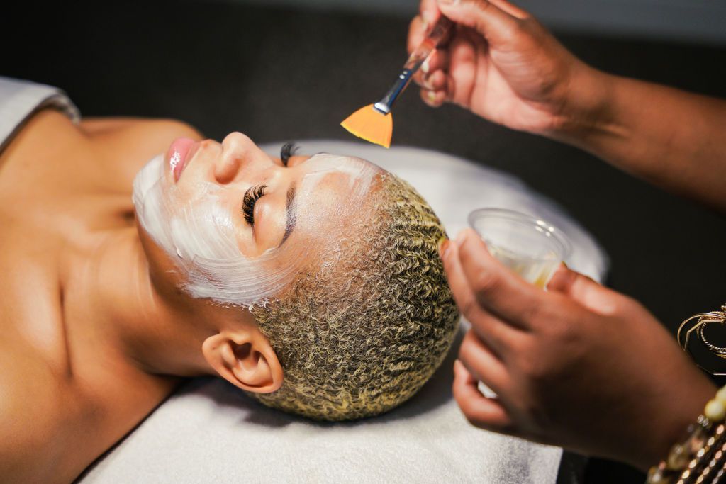 A person with short, light-colored hair lying on a table receives a facial treatment with a brush from an esthetician.