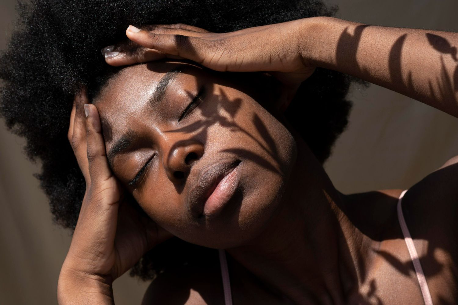 Woman with closed eyes, hands on head, shadows on face, tan background.