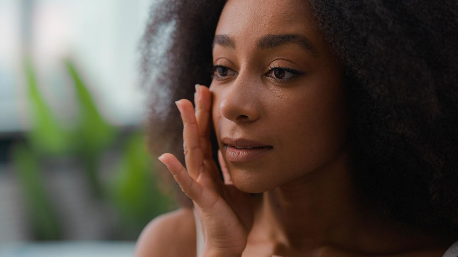 A close-up of a person gently touching their cheek with their fingertips, looking off to the side in a soft, lit setting.