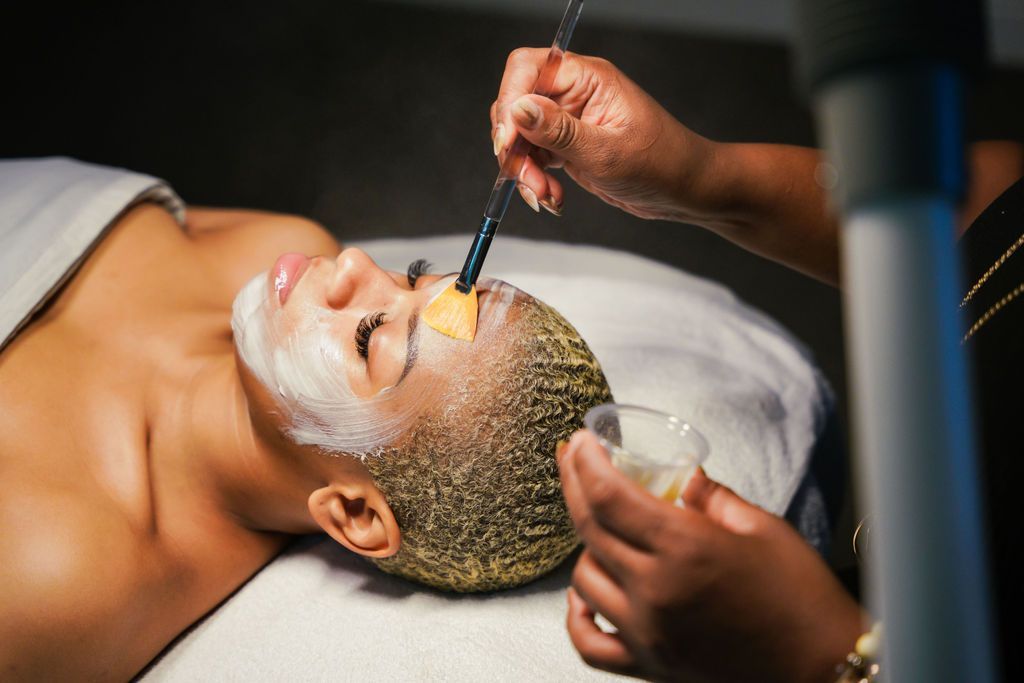 A person with short hair lying on a table while an esthetician applies a white face mask with a brush.