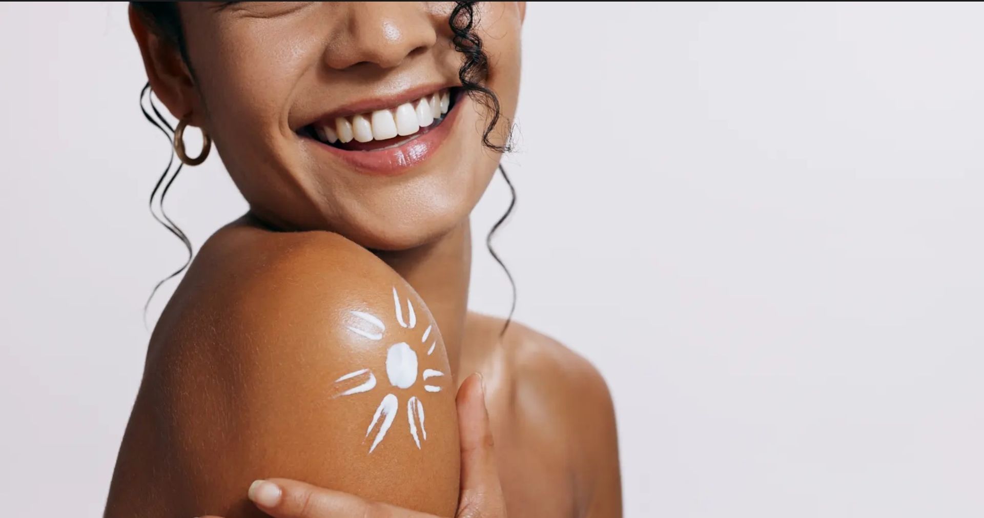 A woman is smiling and applying sunscreen to her shoulder.