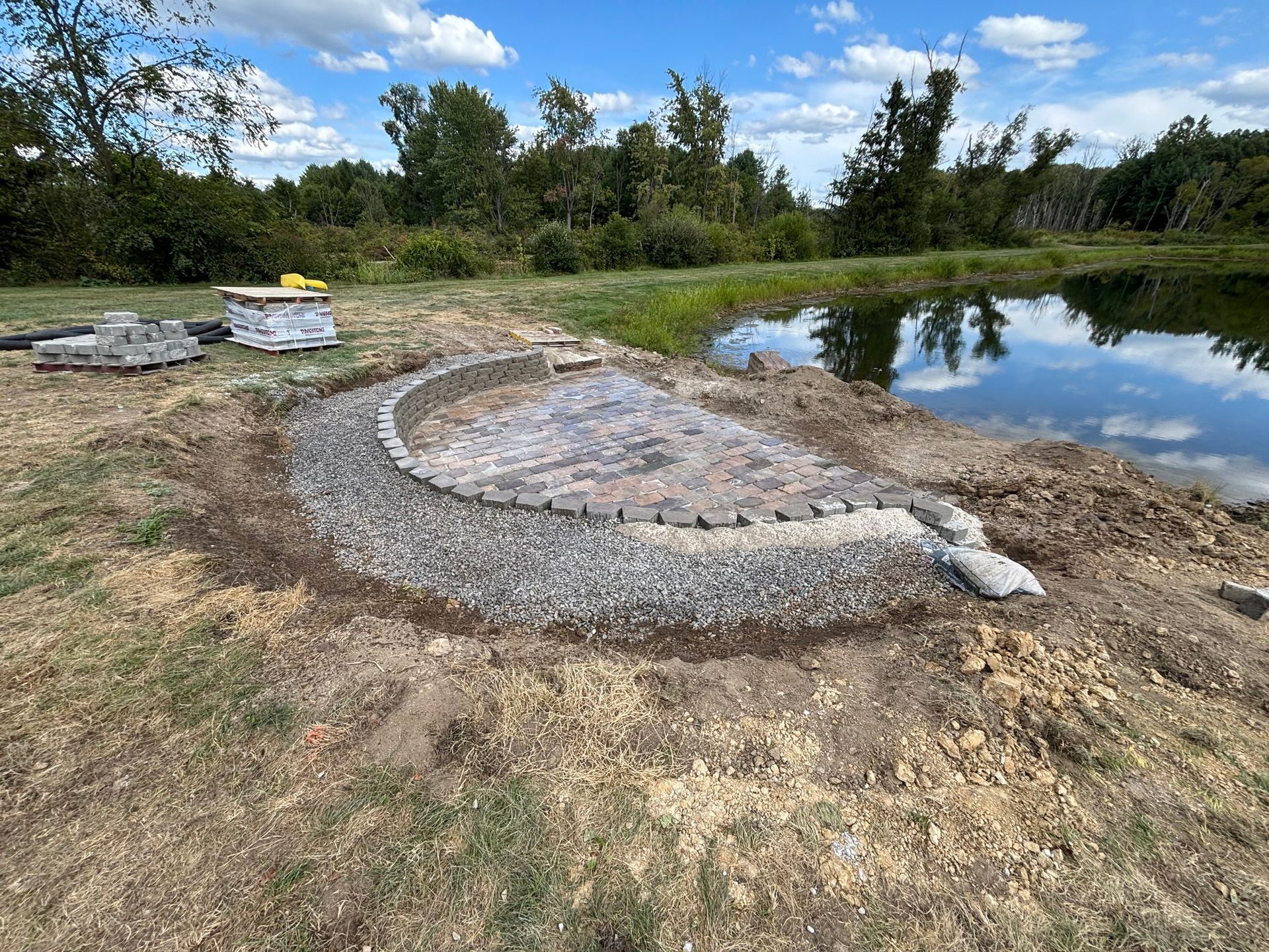A semi-circular brick patio under construction beside a pond, surrounded by gravel, loose pavers, and piles of dirt.