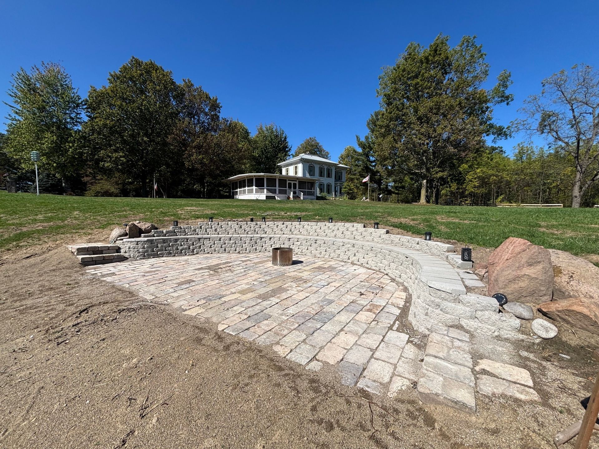A semicircular stone patio with a fire pit sits in a grassy yard, with a white two-story house nestled in the trees behind.