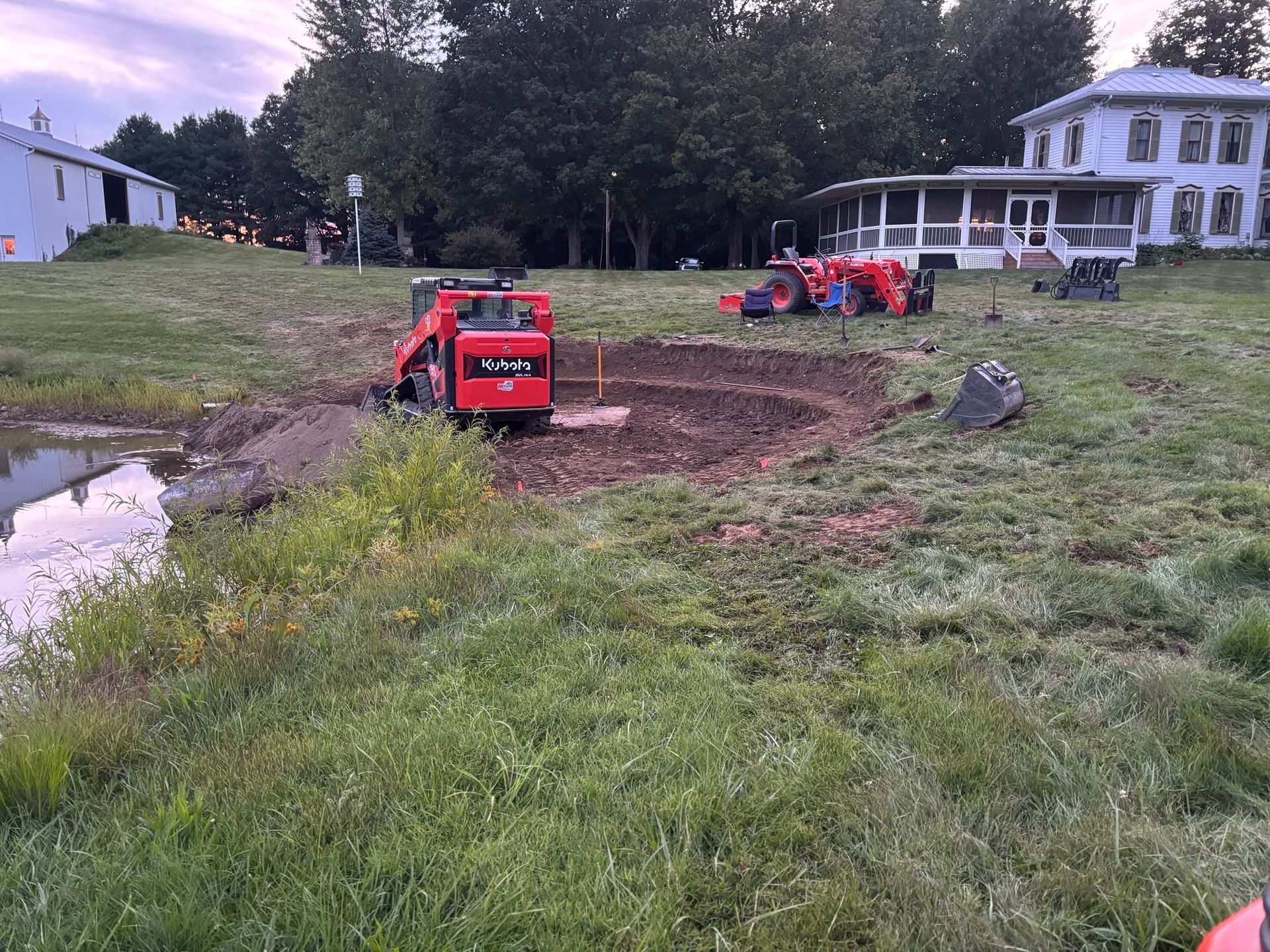 A red skid steer loader performs excavation work near a pond in a grassy yard, with a large house and tractor nearby.