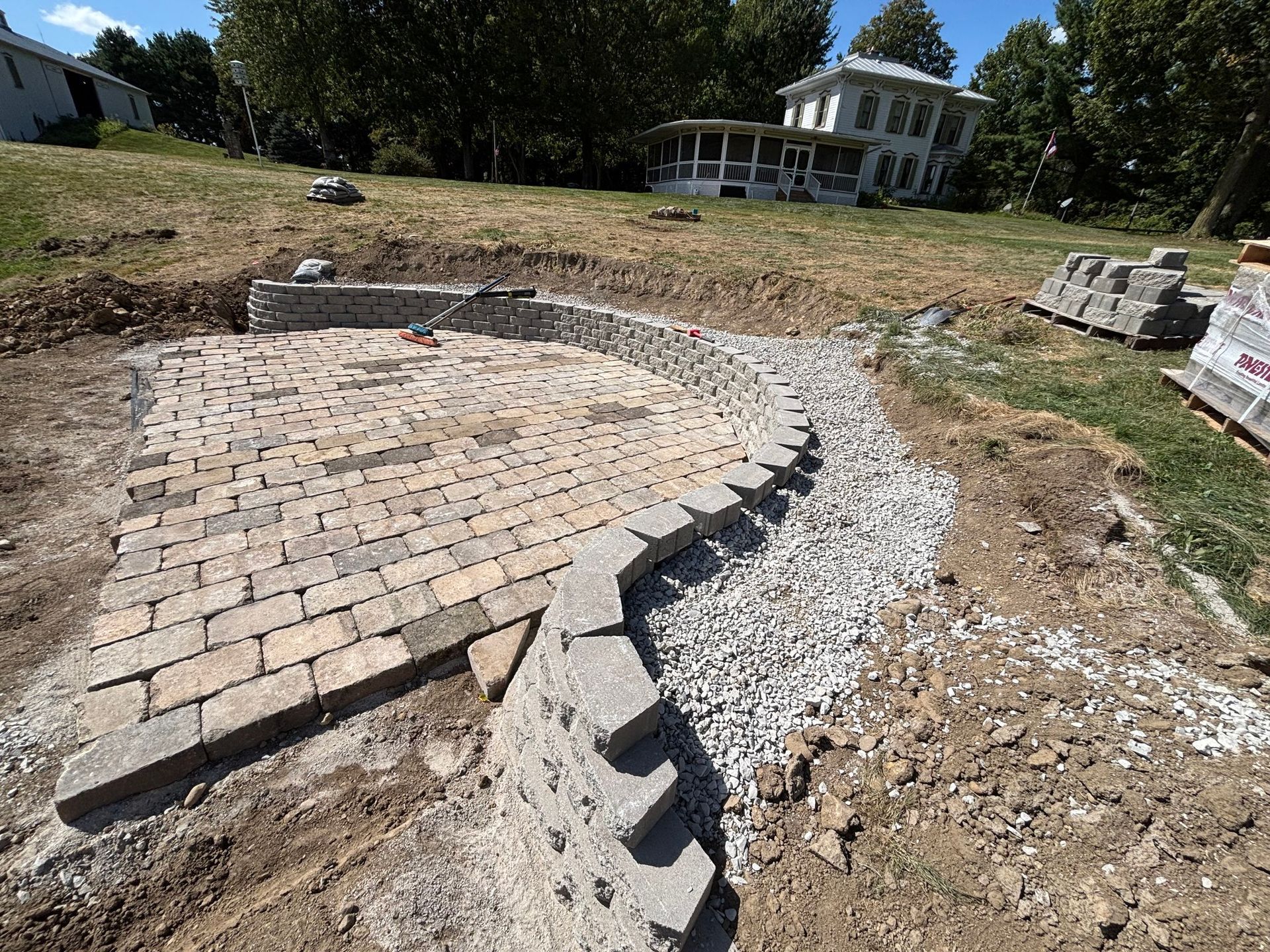 A curved stone retaining wall frames a newly installed paver patio under construction, with gravel and soil nearby.