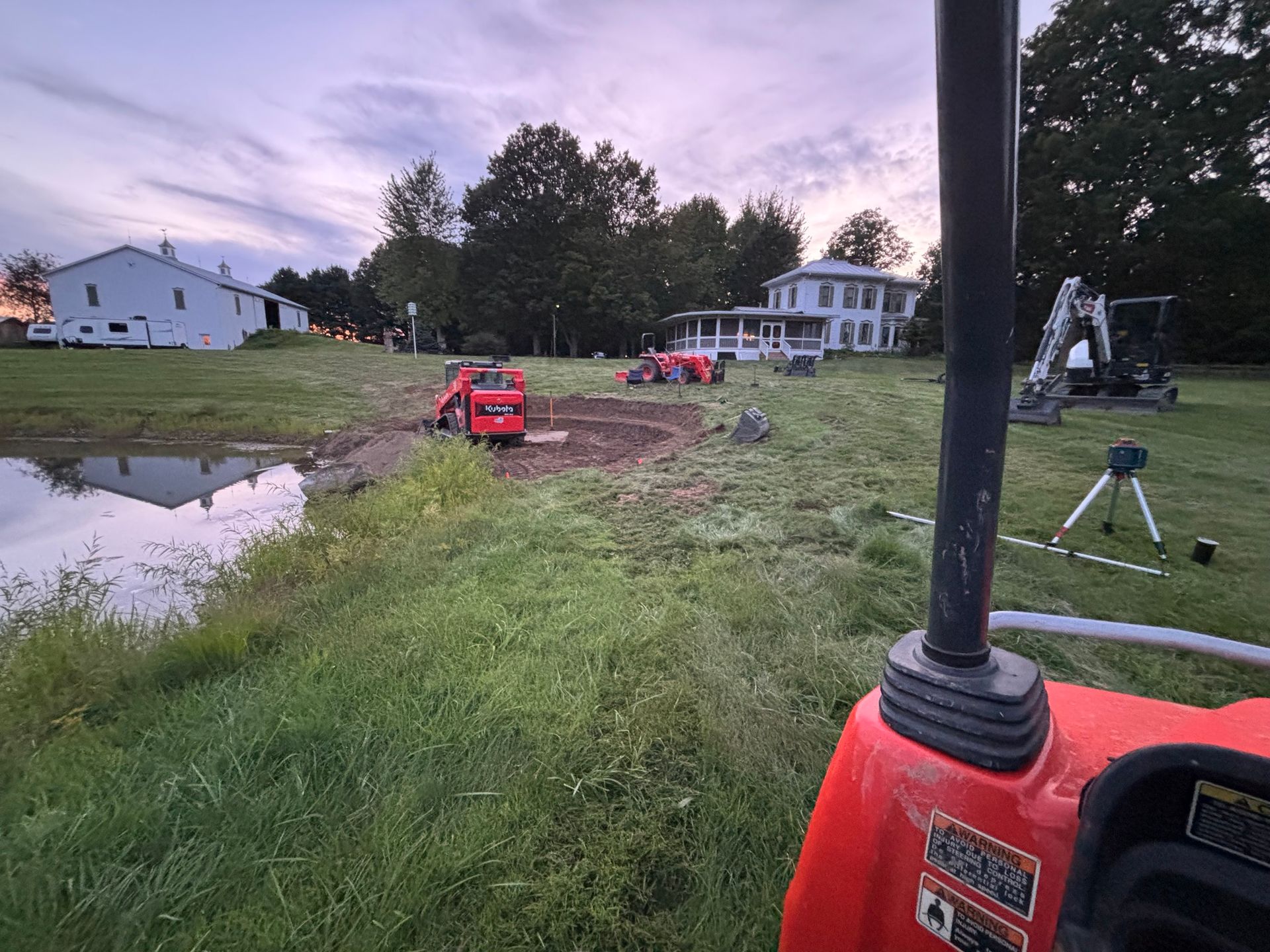 A construction site with two red skid steers and an excavator parked near a pond in front of a white house at dusk.