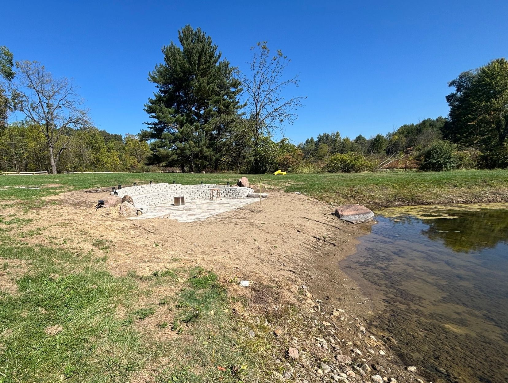 A pile of gray stones on a dirt patch near a pond, set against a grassy field and trees under a clear blue sky.