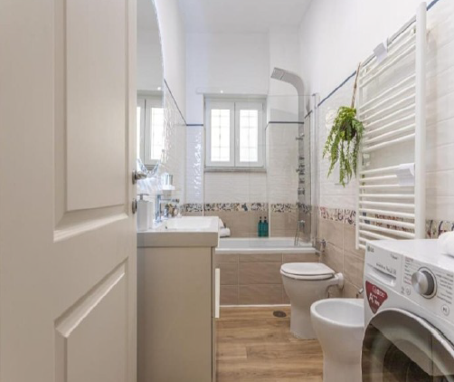 Bagno con porta bianca, lavandino, WC, vasca e lavatrice.