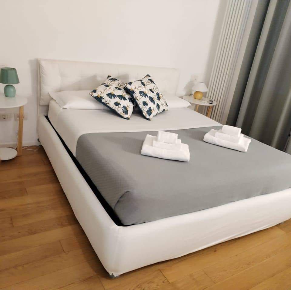Letto bianco con coperta e cuscini grigi in una camera da letto con pavimenti in legno e piccoli tavolini laterali con lampade.