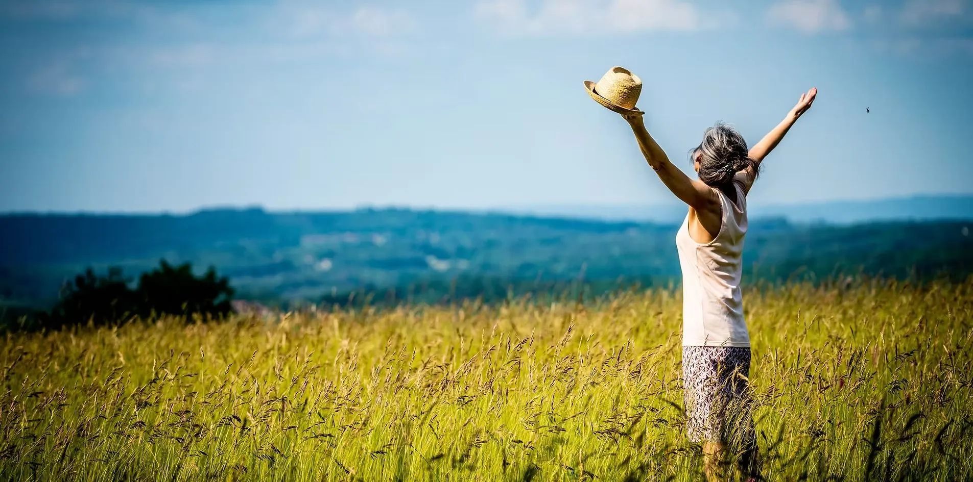 Woman with arms raised, holding a hat, standing in a sunny field, facing distant hills.