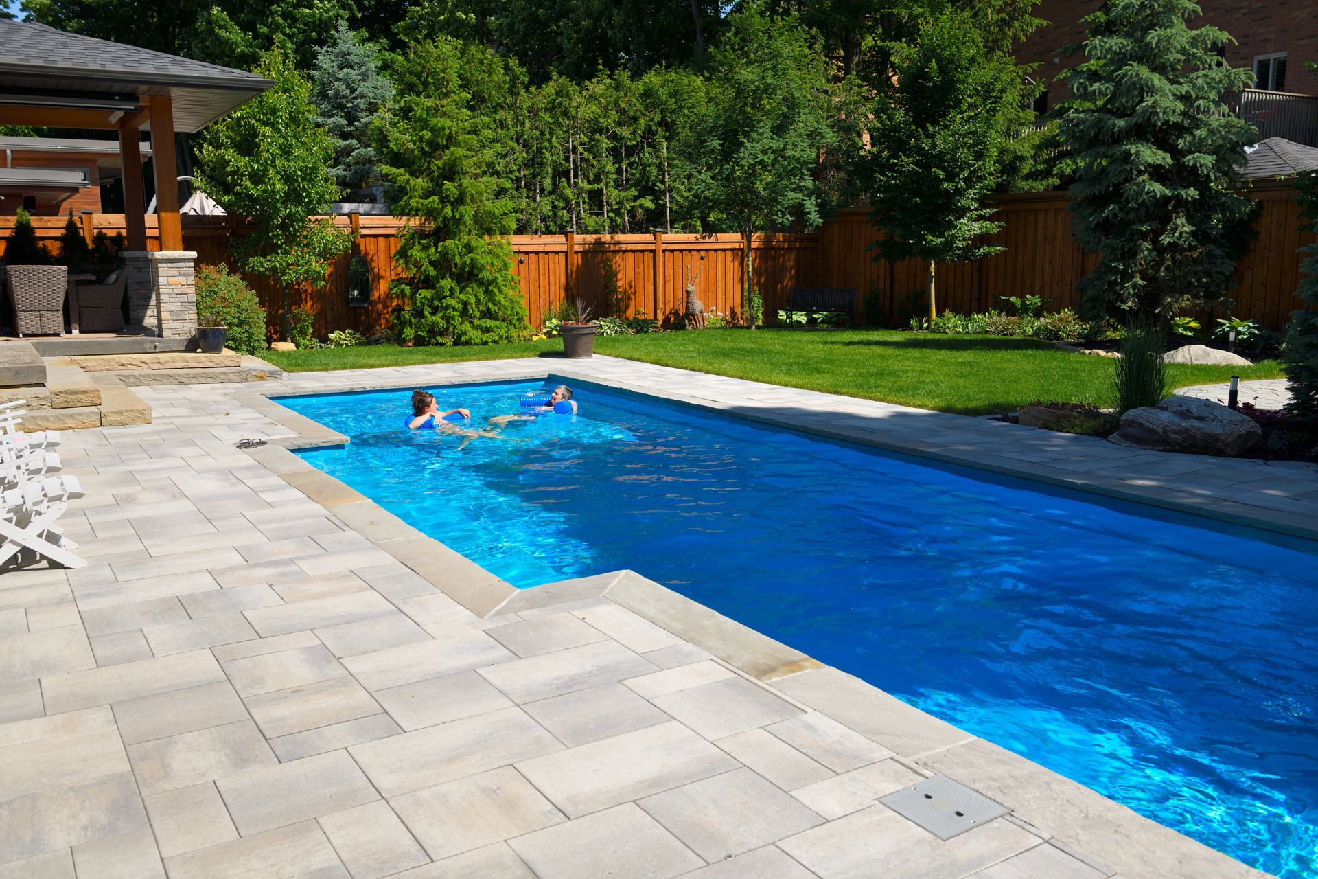 Two women swimming in a new backyard pool with a patio and a green garden