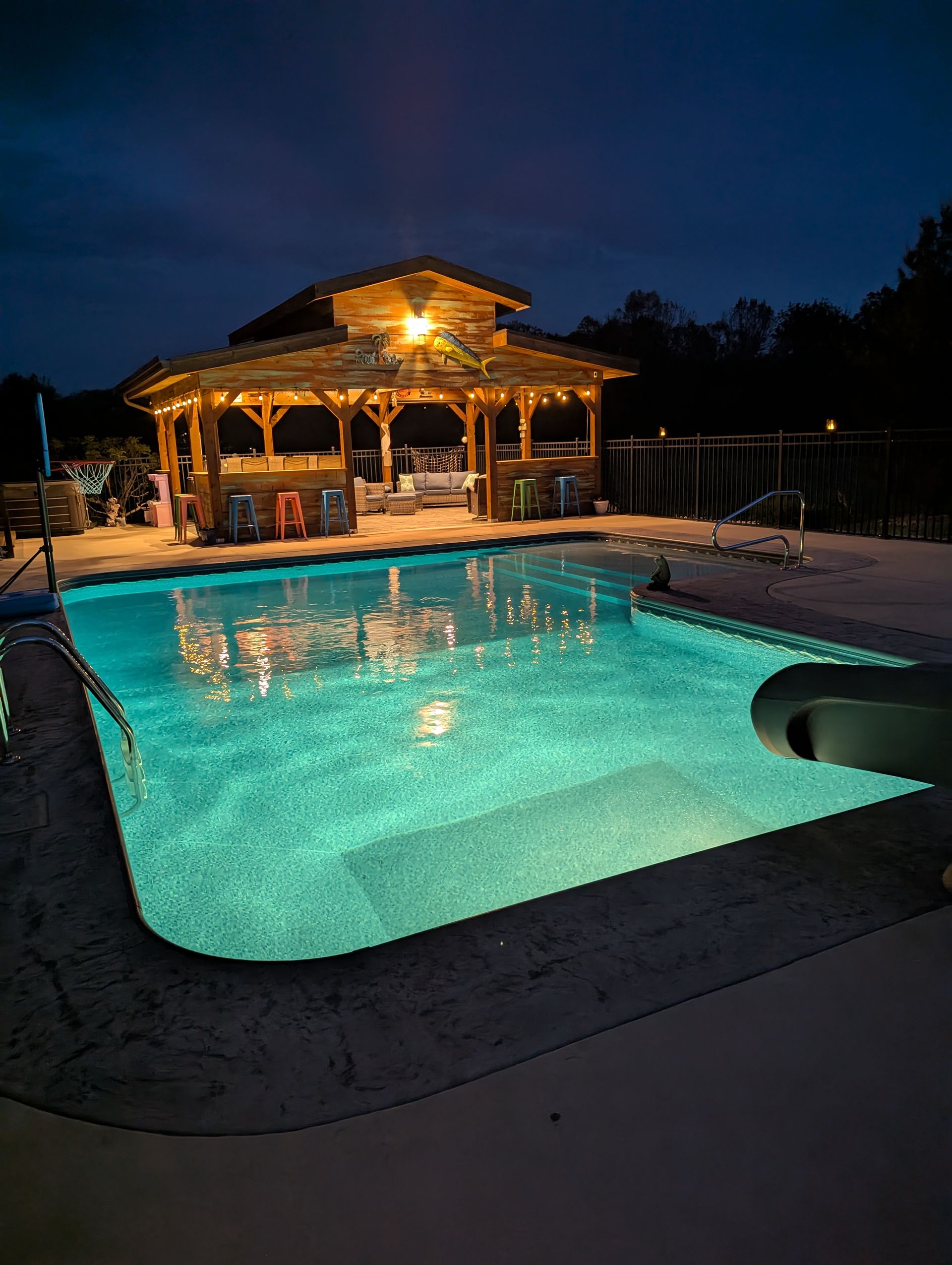 A large swimming pool is lit up at night with a gazebo in the background.
