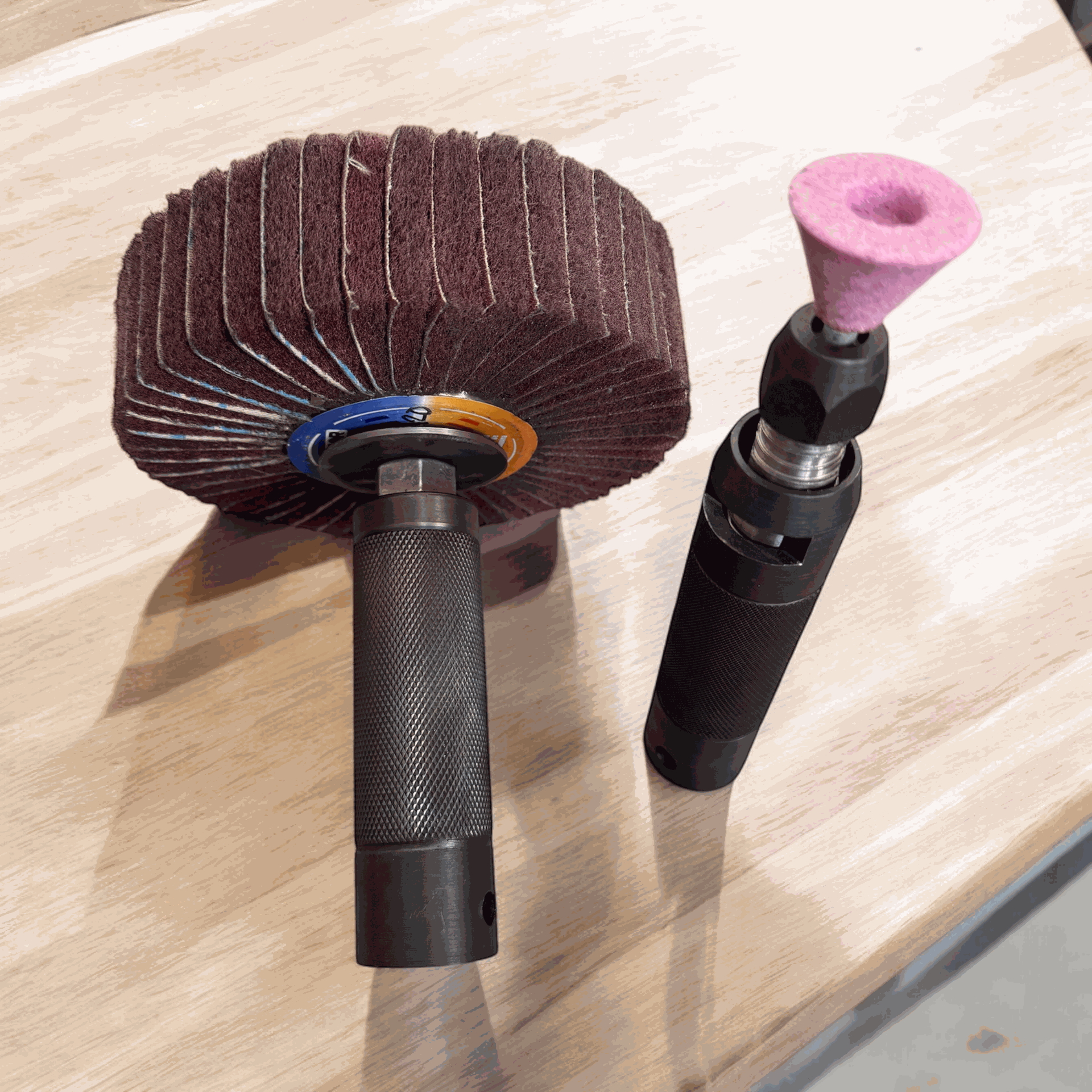 Dos herramientas metálicas con accesorios. Una tiene una rueda abrasiva radial de láminas marrón grande, y la otra, una piedra de afilar cónica rosa.