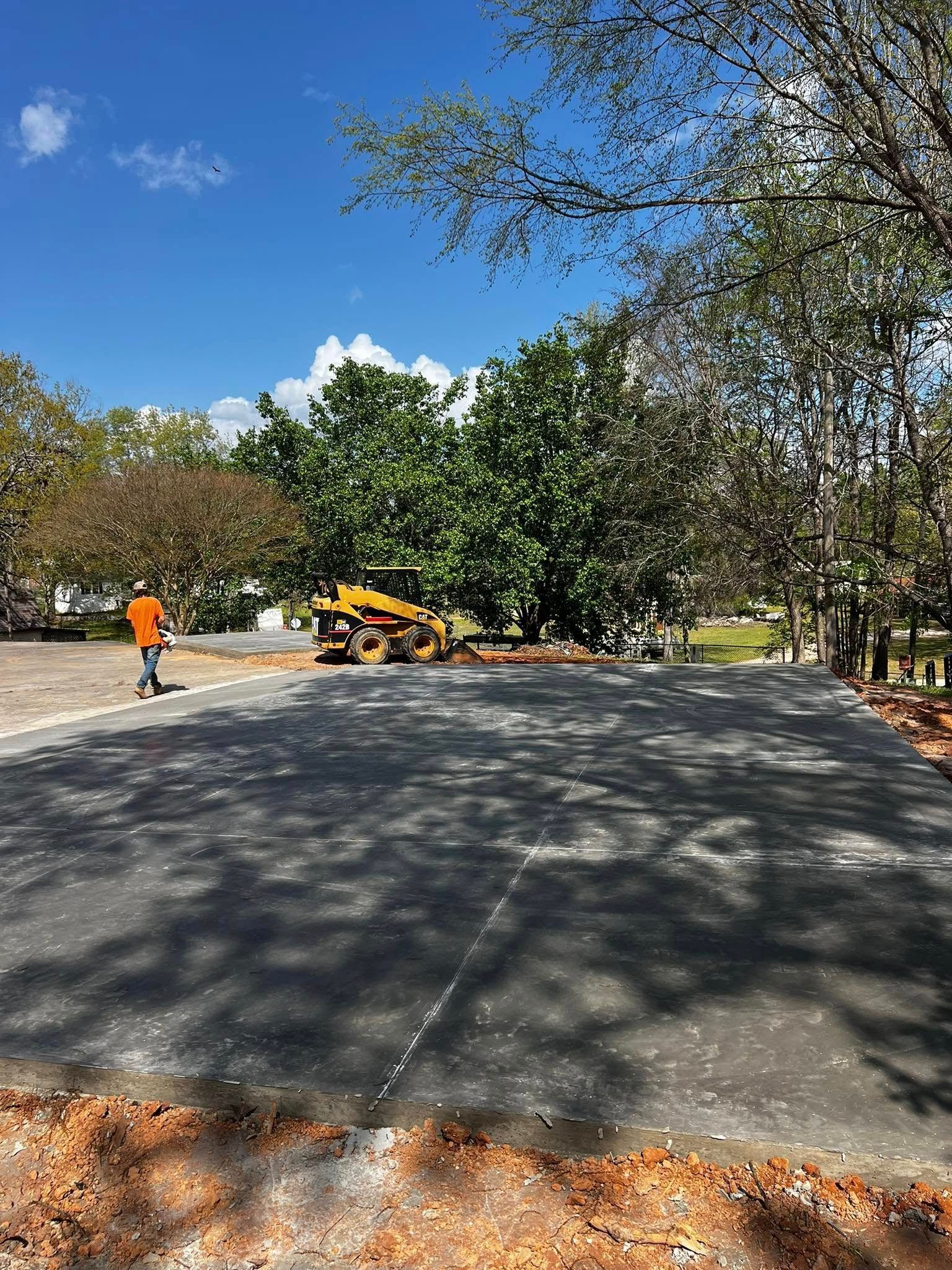 Asphalt paving in progress; worker in orange vest, roller machine, trees, blue sky.
